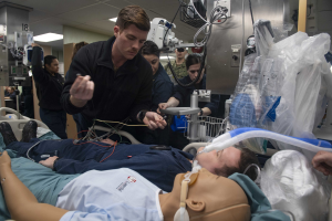 Ein Mann liegt auf einem Krankenhausbett neben einer medizinischen Puppe, umgeben von medizinischem Equipment und Personal in einer klinischen Umgebung.