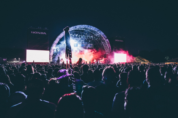Eine große Menge Menschen auf einem Musikfestival mit einem Mann auf einer beleuchteten Bühne umgeben von Musikern, beleuchteten Bildschirmen und Bannern in einer dunklen Umgebung.