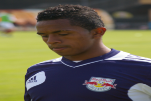 Ein junger Mann in einem blauen T-Shirt steht auf einem Fußballfeld, mit kurzen dunklen Haaren und entschlossener Miene, unscharfer Hintergrund.