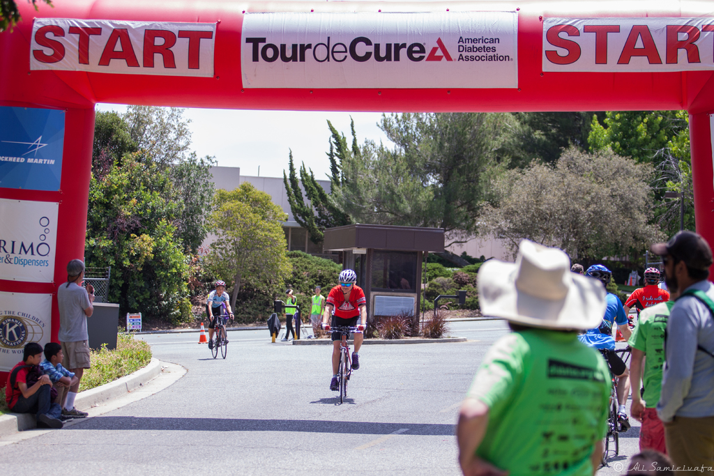 Eine belebte Straßen Szene mit Menschen, die Fahrräder fahren, einige sitzen auf einem Gehweg und andere stehen auf der Straße, mit einer Hütte und umliegenden Pflanzen und Bäumen im Hintergrund.