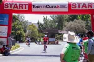 Eine belebte Straßen Szene mit Menschen, die Fahrräder fahren, einige sitzen auf einem Gehweg und andere stehen auf der Straße, mit einer Hütte und umliegenden Pflanzen und Bäumen im Hintergrund.