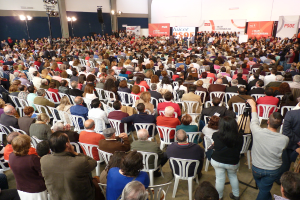 Ein volles Stadion mit vielen sitzenden und stehenden Menschen, eine Bühne mit zwei Rednern am Podium und zahlreiche Banner und Lichter.