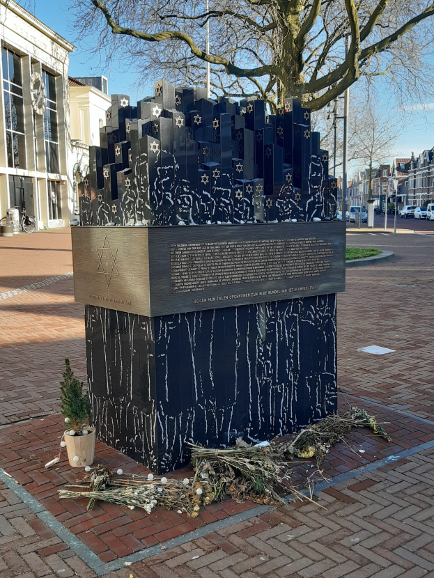 Denkmal in einem Stadtplatz für Holocaust-Opfer, mit einem Baum davor, Topfpflanze, verstreute trockene Blätter, Gebäude, Fahrzeuge, Pfosten, Bäume und Himmel im Hintergrund.