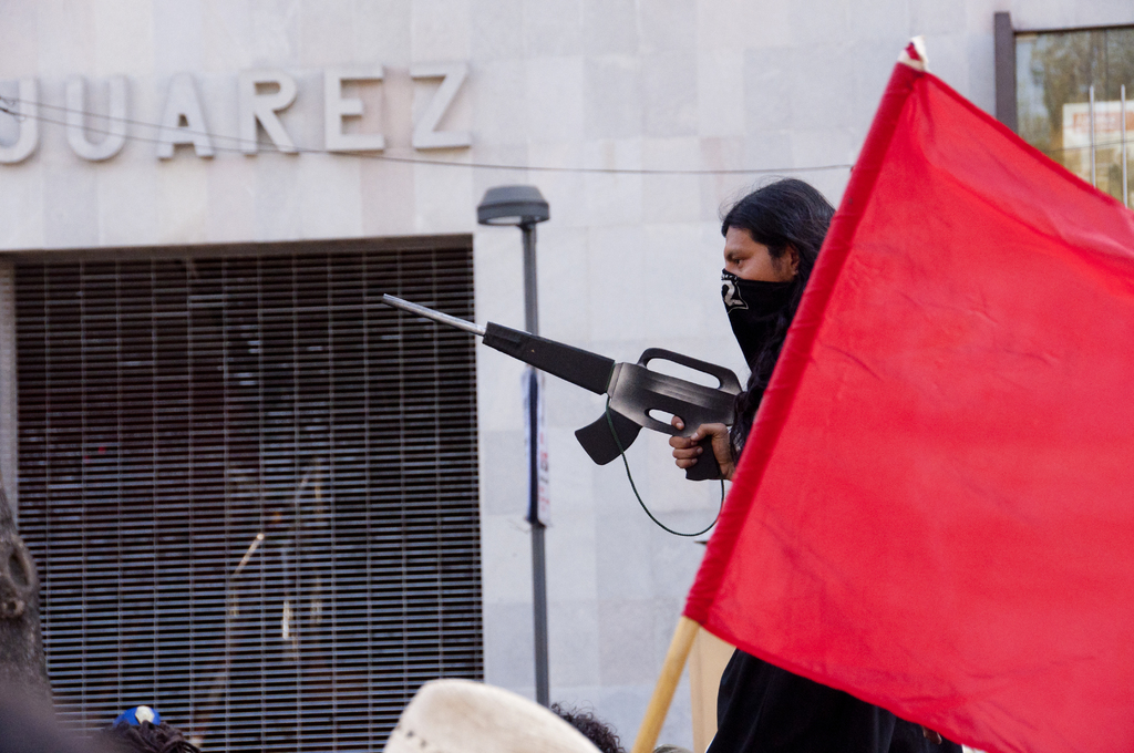 Ein Mann in einem schwarzen Kleid hält eine Waffe und steht neben einer roten Fahne, mit einer Straßenlaterne in der Nähe einer Wand und einem Gebäude im Hintergrund und lesbarem Text in der oberen linken Ecke.