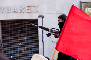 Ein Mann in einem schwarzen Kleid hält eine Waffe und steht neben einer roten Fahne, mit einer Straßenlaterne in der Nähe einer Wand und einem Gebäude im Hintergrund und lesbarem Text in der oberen linken Ecke.