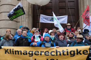 Eine Gruppe von Menschen hält ein Transparent, Fahnen und Luftballons vor einem Gebäude mit Säulen und einer Tür während eines Klimastreiks in Deutschland.