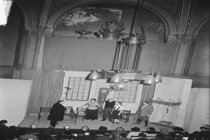 Eine Gruppe von Menschen auf einer Bühne vor einem sitzenden Publikum in einem Raum mit Kronleuchtern, Vorhängen, Fenstern und einer Wand im Hintergrund, dargestellt in Schwarz-Weiß.