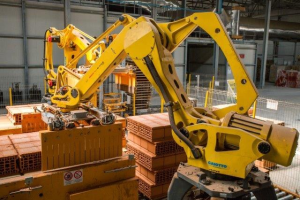 Großes industrielles Lagerhaus mit gestapelten Kisten, Überkopfkranen, Zaun, Strommasten und einer fernen Wand in einer automatisierten Fertigungsumgebung.