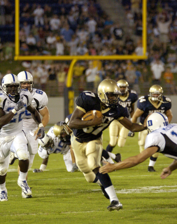 Ein Fußballspieler in Bewegung mit dem Ball, umgeben von Gegenspielern und Teamkollegen in Sportkleidung und Helmen, mit einem unscharfen Stadionhintergrund und Text unten.