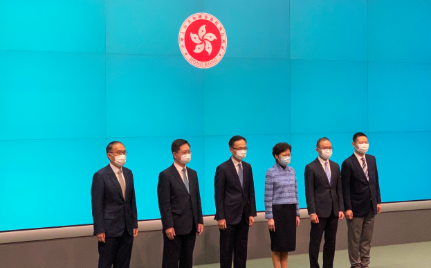 Eine Gruppe maskierter Individuals stehend nebeneinander vor einer blauen Wand mit einem Logo, an einer Pressekonferenz in Hong Kong teilnehmend.