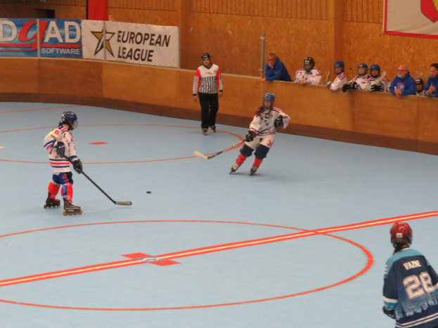 Indoor-Eishockeyspiel mit Spielern in Helmen und Schlittschuhen, die Stöcke halten, Wandbannern und Zuschauern in den Rängen.