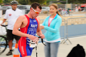 Ein Mann in einem roten und blauen Triathlonanzug läuft eine Straße entlang, neben ihm geht eine Frau, während im Hintergrund Menschen, Fahrräder, Verkehrskegel, Geländer, Fahrzeuge, Bäume und andere Gegenstände zu sehen sind.