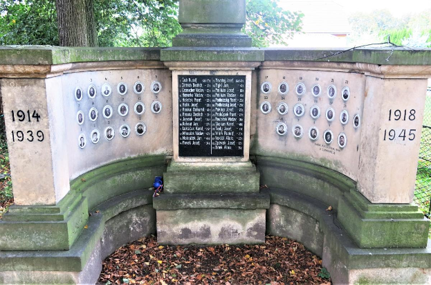 Ein Denkmal in einem von Bäumen und einem Zaun umgebenen Park mit einer Tafel an der Wand und trockenen Blättern auf dem Boden, das sich auf dem jüdischen Friedhof in Berlin, Deutschland befindet und als Gedenkstätte für Holocaust-Opfer dient.