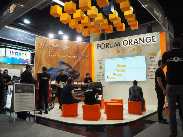 Eine Gruppe von Menschen an einem IT-Cube Systems Forum Orange Cyber Security Stand, einige sitzen, andere stehen, umgeben von einem Bildschirm, Regalen mit Gegenständen, Texttafeln, Deckenleuchten und Lampen.