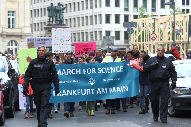 Eine Gruppe von Menschen marschiert mit einer 'March for Science Frankfurt am Main'-Fahne die Straße entlang, Autos, Gebäude, Statuen, Laternen, Schilder und Bäume im Hintergrund.