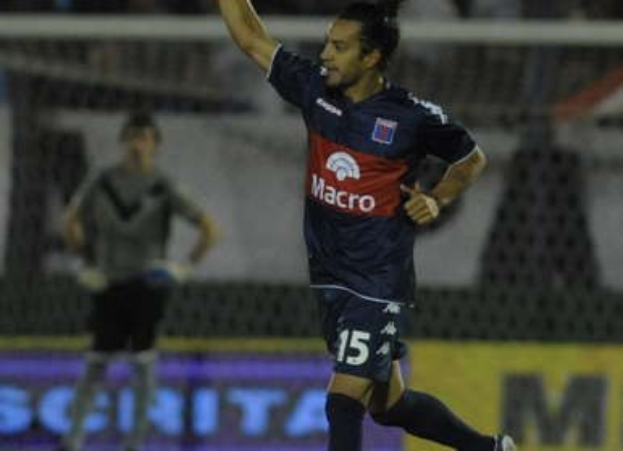 Soccer player on the ground celebrating a goal, with a blurred crowd and a person standing in the background.