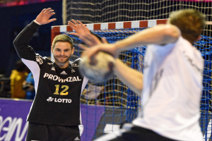 Zwei Männer spielen Handball vor einem Tor mit Netz und einer Fahne in einer Stadionatmosphäre.