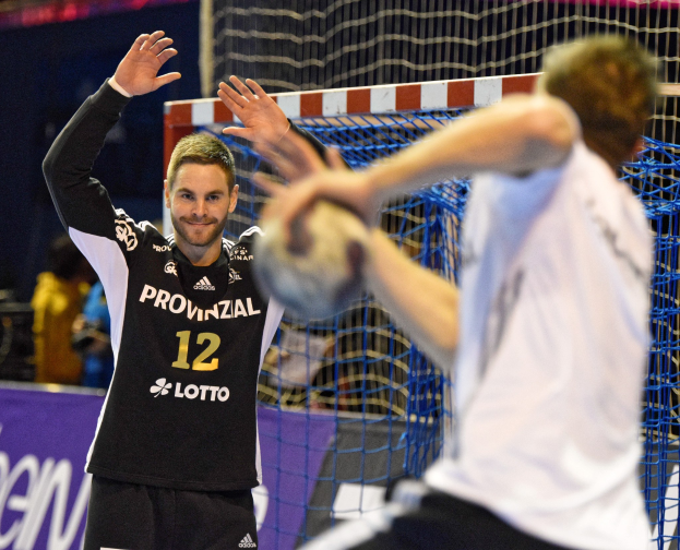 Zwei Männer spielen Handball vor einem Tor mit Netz und einer Fahne in einer Stadionatmosphäre.
