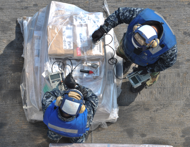 Zwei Männer in blauen Uniformen und Helmen arbeiten an einem großen Drohnenmodell, halten verschiedene Gegenstände und haben eine Plastikabdeckung vor sich auf dem Boden liegen.