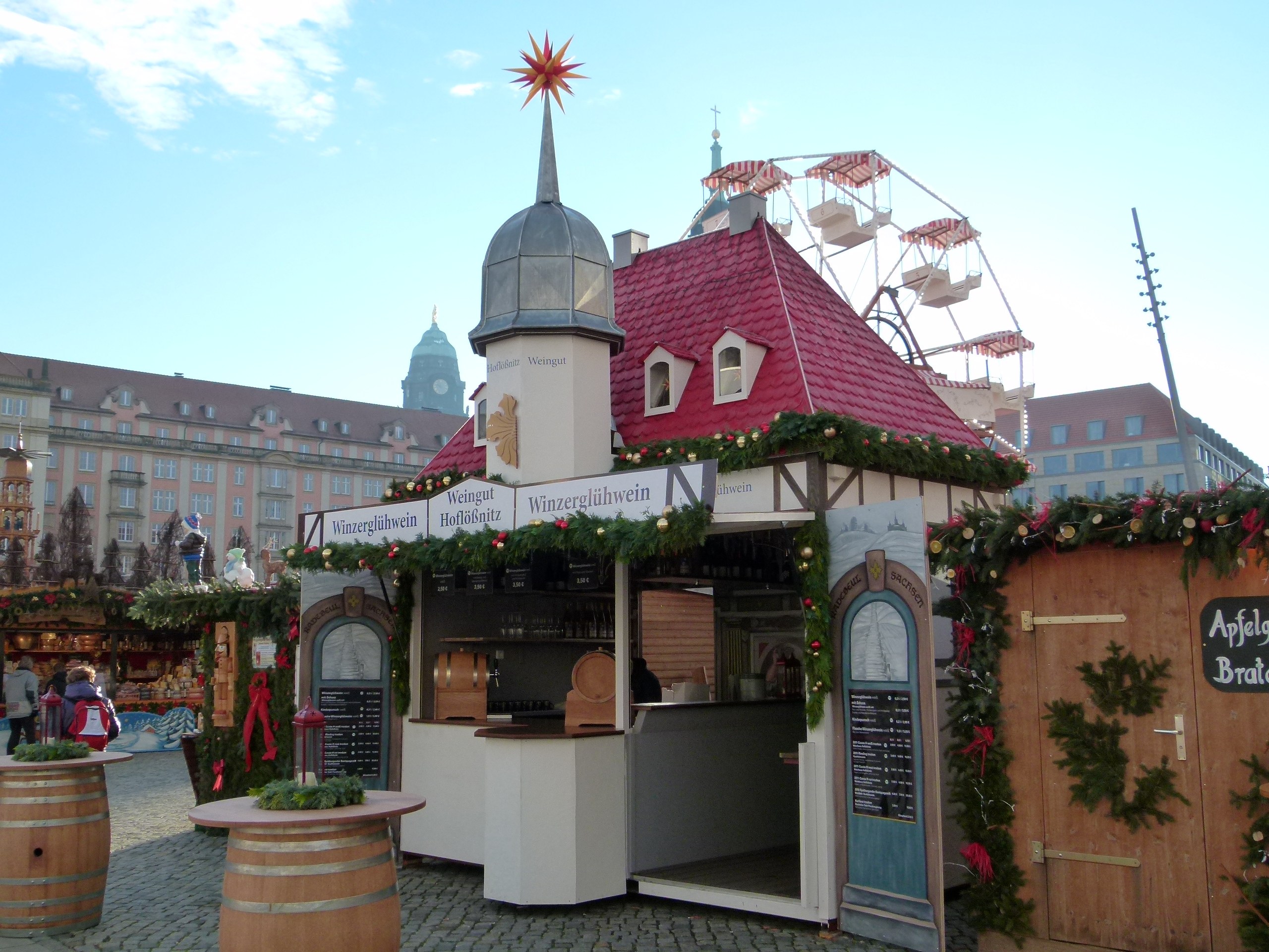 Ein kleines rotes Gebäude mit einem Riesenrad davor, umgeben von Festdekorationen und Menschen, vor einer Kulisse aus Gebäuden, Bäumen und einem bewölkten Himmel während des Oktoberfests in München, Deutschland.