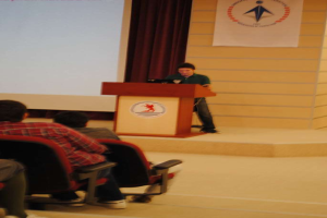 Eine Person steht auf einem Podium in der Mitte, spricht, mit mehreren sitzenden Individuen links, eine Ebene tiefer, und Bretter und ein Vorhang auf der linken Seite.