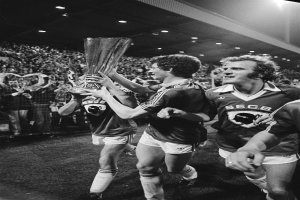 Schwarzes und weißes Bild von Fussballspielern, die mit einem Pokal auf einem Feld feiern, umgeben von einer Menge hinter einem Zaun unter Stadionbeleuchtung.