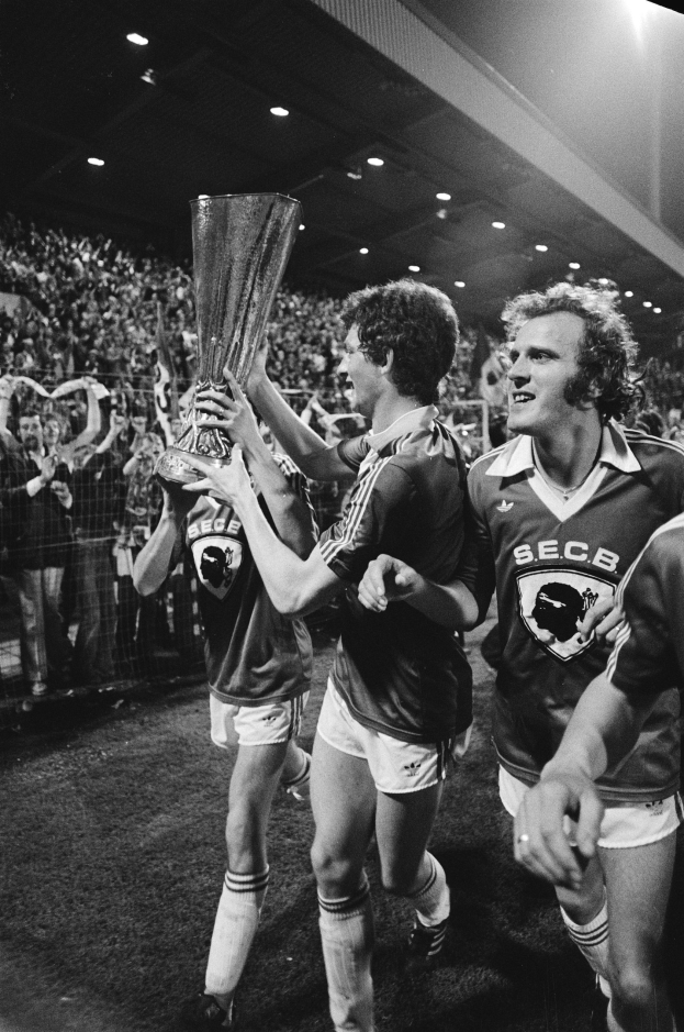 Schwarzes und weißes Bild von Fussballspielern, die mit einem Pokal auf einem Feld feiern, umgeben von einer Menge hinter einem Zaun unter Stadionbeleuchtung.