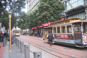 Ein Zug auf Eisenbahnschienen mit Text an der Seite, eine Person in der Nähe, kleine Pfosten mit Seilen auf der linken Seite, Straßenlaternen, Bäume, viele Menschen, ein Müllcontainer auf der linken Seite und Gebäude mit Leitern auf der rechten Seite.
