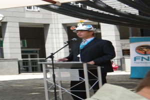 Ein Mann in Anzug und Hut spricht in ein Mikrofon an einem Podium, mit einer Person rechts unten, einem Banner mit Text rechts und einem Gebäude mit Geländer im Hintergrund.