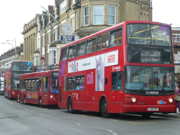 Eine Gruppe roter Doppeldeckerbusse fährt eine Straße mit hohen Gebäuden, Fußgängern auf dem Gehweg, Laternen und einem Gebäudeuhr an der Wand entlang.