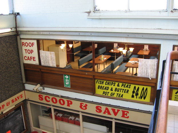 Restaurant-Innenraum mit Tischen, Stühlen und beleuchteten Tafeln mit Text, mit einem Geländer auf der rechten Seite und einer Wand im Hintergrund, mit einem "Scoop & Save"-Café-Schild.