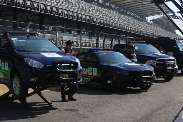 Eine Gruppe von Polizeiwagen vor einer Rennstrecke geparkt, uniformierte Personen auf der Straße stehend, ein Stadion mit Geländern und Maschendrahtzaun im Hintergrund und der Himmel oben sichtbar.