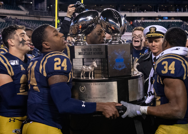 Eine Gruppe von Männern hält einen Pokal vor einer jubelnden Menge in einem hell erleuchteten Stadion, wobei der Mann in der Mitte eine Kamera hält.