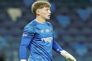 Ein junger Mann in einem blauen Torwarttrikot und weißen Handschuhen steht auf einem Fußballfeld und scheint mitten in einem Spiel zu sein, mit einem unscharfen Hintergrund.