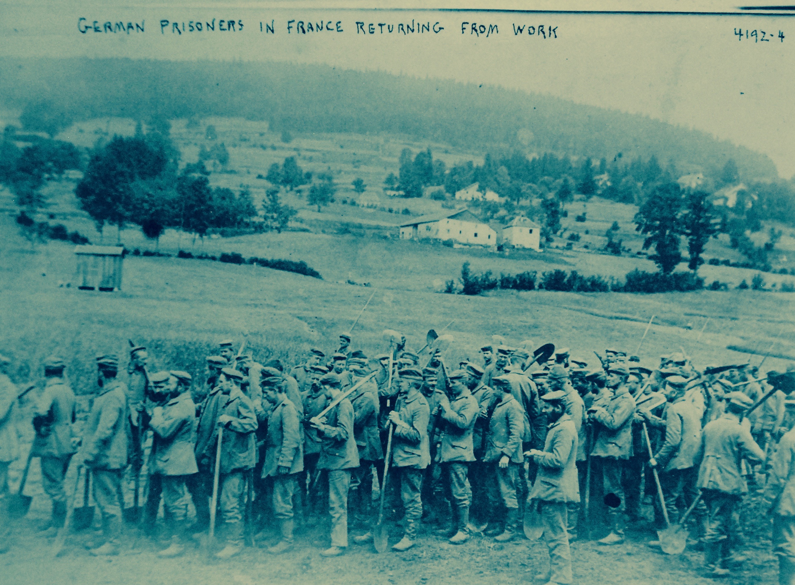 Eine Gruppe deutscher Gefangener in Frankreich, die Gewehre in den Händen halten, steht auf dem Boden mit Bäumen, Häusern und einem klaren blauen Himmel im Hintergrund.