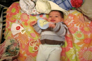 Ein Baby liegt auf einem Bett mit Spielzeugen, Kissen und Kleidung daneben.