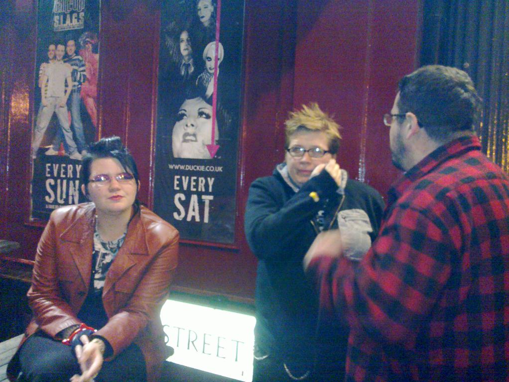 Eine Frau sitzt auf einer Bank mit zwei Männern neben ihr, vor einer Wand mit Filmplakaten.