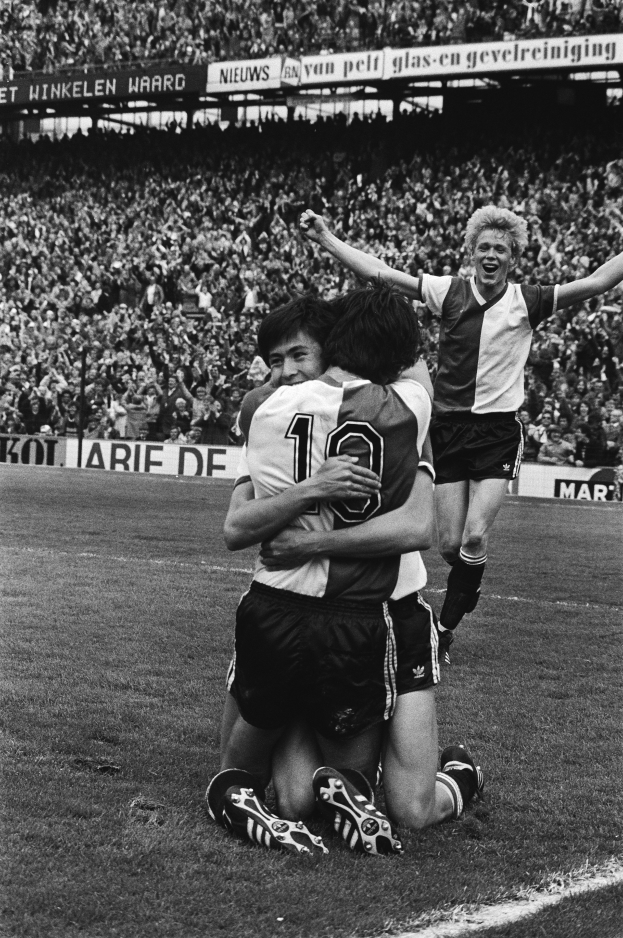 Drei Fussballspieler feiern ein Tor auf dem Boden vor einer jubelnden Menge in einer schwarz-weißen Stadionszene.