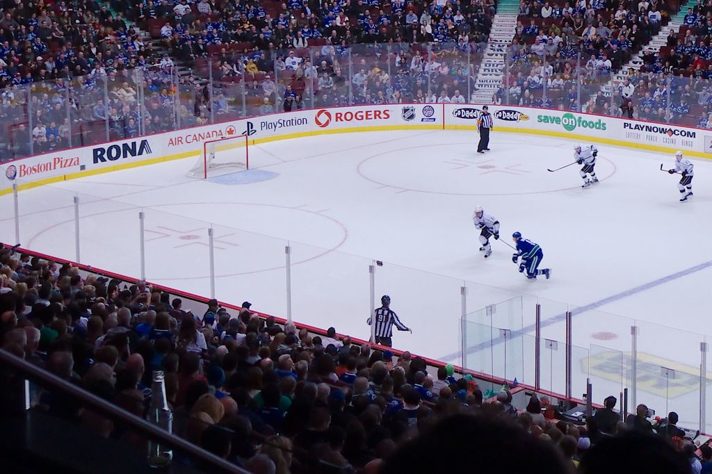 Vier Männer spielen Hockey auf einem Eisstadion, zwei stehen in der Nähe des Tors und des Zauns, Zuschauer sitzen auf Stühlen im Hintergrund und eine Treppe ist sichtbar.