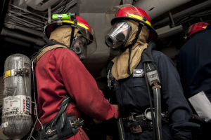 Zwei Feuerwehrleute in Helmen und Gasmasken stehen neben einem Sauerstoffzylinder, mit Rohren und anderen Gegenständen im Hintergrund.