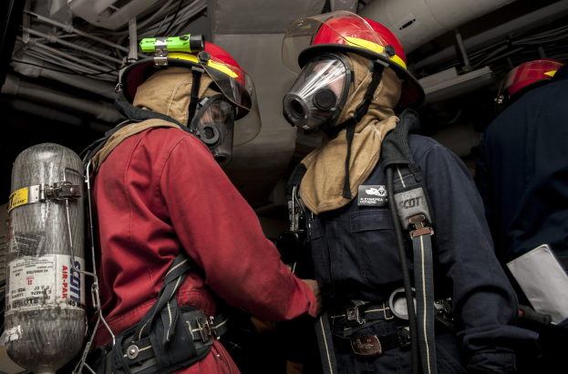Zwei Feuerwehrleute in Helmen und Gasmasken stehen neben einem Sauerstoffzylinder, mit Rohren und anderen Gegenständen im Hintergrund.