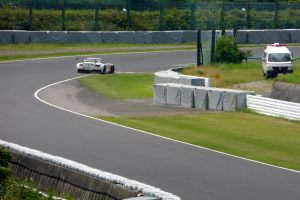 Ein Rennwagen fährt eine Rennstrecke hinunter mit einem Polizeiwagen im Hintergrund, umgeben von Gras, Pflanzen, Bäumen und Absperrungen, mit einem Fahrzeug auf der rechten Seite und Text unten.
