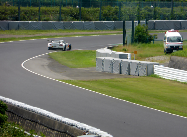 Ein Rennwagen fährt eine Rennstrecke hinunter mit einem Polizeiwagen im Hintergrund, umgeben von Gras, Pflanzen, Bäumen und Absperrungen, mit einem Fahrzeug auf der rechten Seite und Text unten.