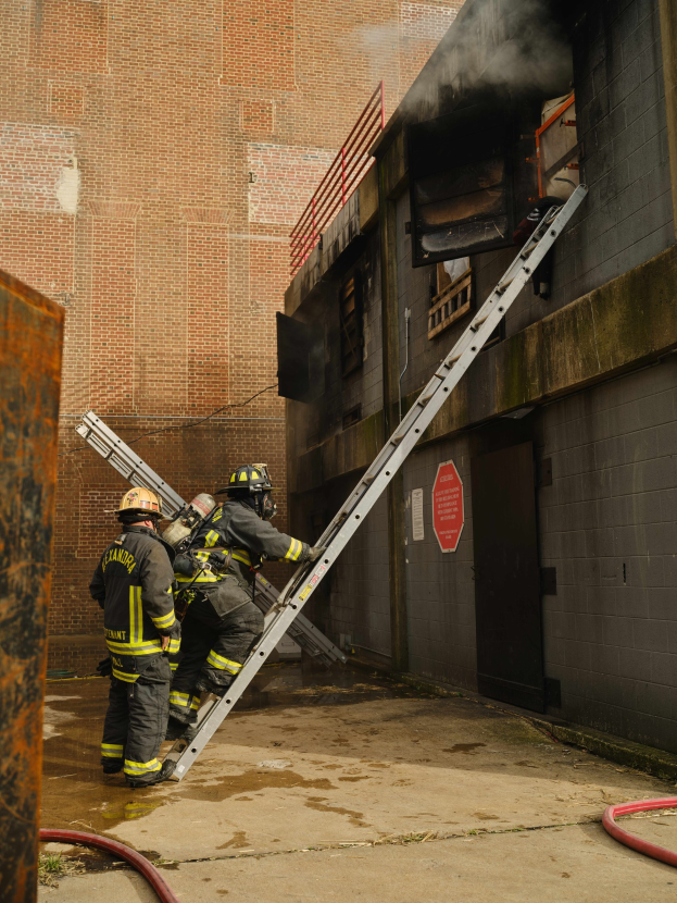 Feuerwehrleute mit Helmen klettern an einer Leiter zu einem raucherfüllten Gebäude hinauf, mit einem Metallobjekt links und an der Unterseite sichtbaren Rohren.
