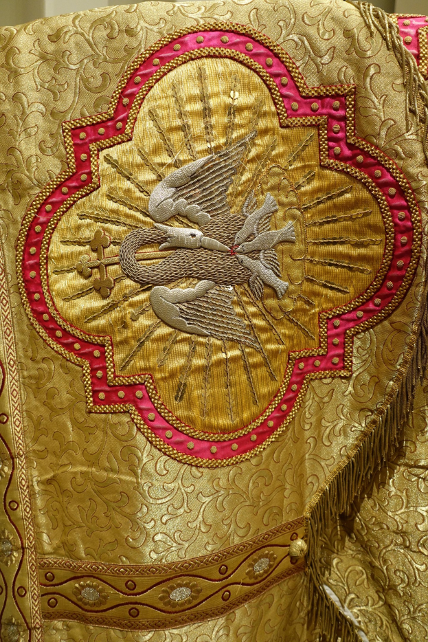 Nahaufnahme eines Priestergewandes mit goldenen und roten Stickereien auf einem reichhaltigen Stoff, vor einer neutralen Wand.