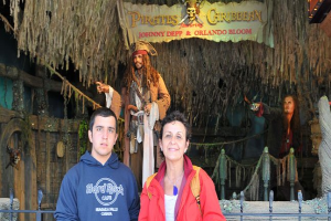 Ein Mann und eine Frau stehen vor einem Piratenschiff, mit Statuen von kostümierten Figuren, einem Banner mit Text und Lichtern im Hintergrund und einem Geländer am unteren Bildrand.