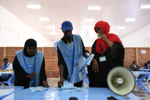 Eine Gruppe von Menschen steht um einen Tisch mit einem Megafon und Papieren, während andere im Hintergrund unter einer beleuchteten Decke sitzen oder stehen.