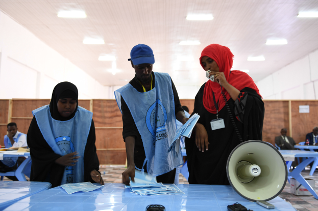 Eine Gruppe von Menschen steht um einen Tisch mit einem Megafon und Papieren, während andere im Hintergrund unter einer beleuchteten Decke sitzen oder stehen.