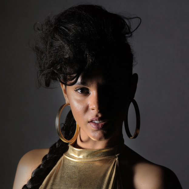 Eine Frau in einem goldenen Kleid und großen Creolen steht selbstbewusst vor einer Wand, ihr glatter Bob-Haarschnitt und das natürliche Make-up betonen ihren entschlossenen Gesichtsausdruck, während sie direkt in die Kamera schaut.