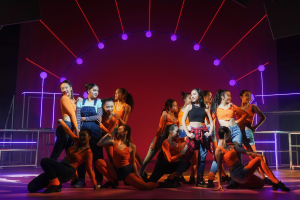 Eine Gruppe junger Tänzer in farbenfrohen Kostümen performs auf einer Bühne mit Geländern, die Arme erhoben, während sie von bunten Lichtern und gemusterten Dekorationen umgeben sind und ein applaudierendes Publikum zusieht.